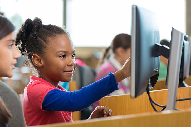 Elementary age African American little girl is smiling while using a desktop computer in public school library or computer lab. Diverse students are wearing casual clothing. Child is pointing at computer monitor screen while working on school assignment with friend.